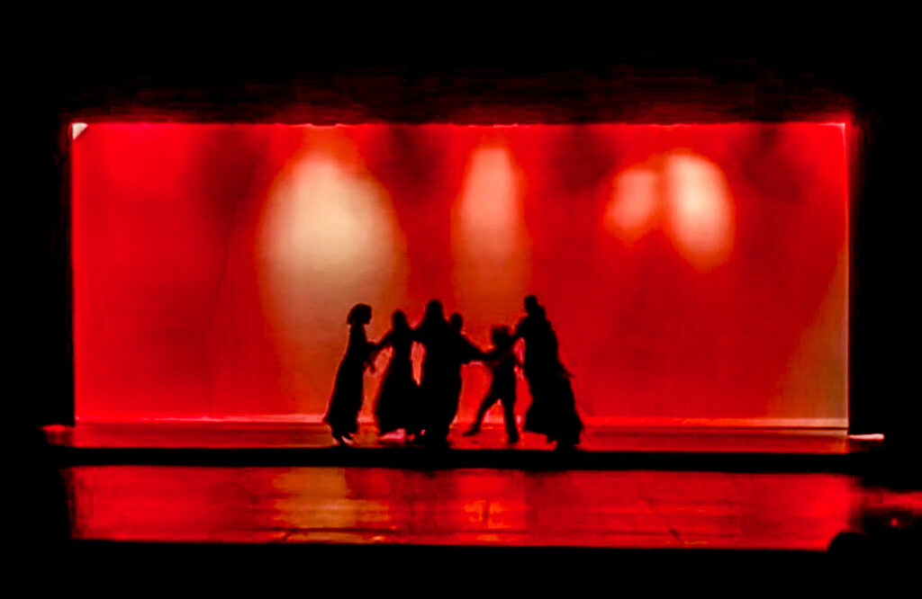 In silhouette against a red backdrop, girls dance in a circle.